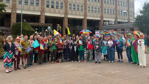 Henry Durand/Local Democracy Reporting Service A group of people gather outside County Hall in Norfolk, holding up various rainbow-coloured symbols, flags and placards