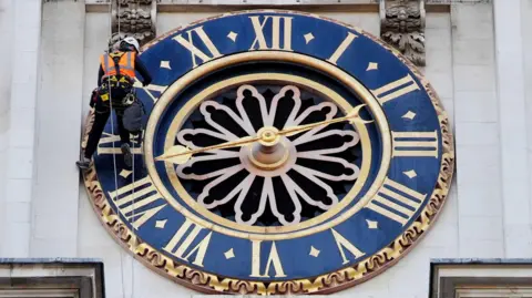 PA Media A man in high-visibility wear surveys a blue and gold clock face on the Hawksmoor clock tower at Westminster Abbey.