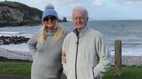 A woman in a blue hat and grey sweater linking arms with a man with grey hair in a cream zip up. They are standing in front of a pebbled beach with rocks in the background. 