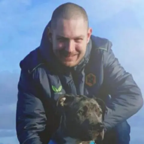 Family A man with cropped brown hair, wearing a blue coat with an orange Wolves FC logo on the chest. He is crouched down with a black dog in front of him.