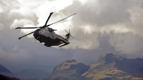 An RAF helicopter above a mountain range - it flies low to the cliff face against a dramatic grey cloud.