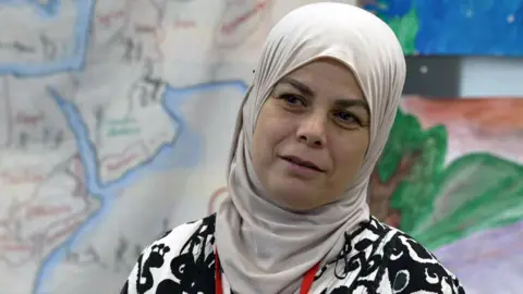 Warda is wearing a grey hijab around her head and neck and a black and white monochrome top. She is standing in front of a very large painting depicting a map.