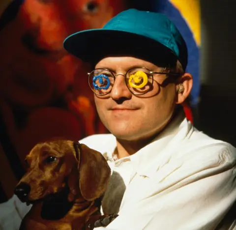 Getty Images David Hockney is holding a brown dog and is wearing a blueish green baseball cap and glasses with spiral designs on them. 