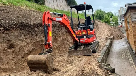 A mini-digger is parked on a muddy track with a muddy bank to its left and a dropped walkway to the right. There is no driver. The bucket is resting on the mud. There is a grassy bank and wooden fence above it and a shed to the right. 