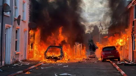 PA Media Large orange flames engulf a residential street. Houses and cars are on fire as black smoke billows, obscuring almost everything. One car, on the left, is almost entirely burnt out.