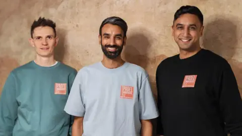 The trio are wearing light coloured T-shirts or tops with their branding on - an orange square with Padel Warehouse 52. They are smiling