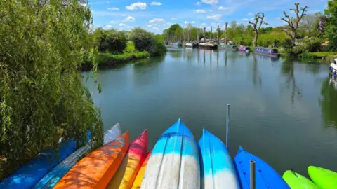 BBC Weather Watcher/YorkshireTed Several different-coloured canoes lying upside down in the foreground at the edge of a canal which stretches ahead in the sunshine. The sky is blue with white fluffy clouds, the banks of the canal have trees and shrubs and there are several moored canalboats on the right.