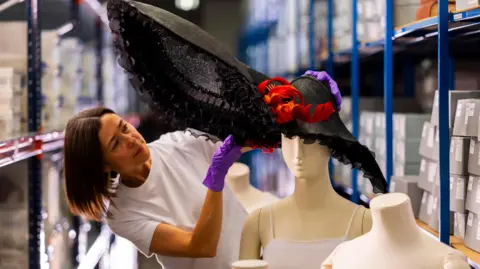 A woman wearing a white blank t-shirt and purple gloves is angling a large hat that is black with red feathers atop a mannequin
