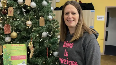 Nicky is pictured wearing a grey hoody with pink Time is Precious writing with a Christmas tree in the background. It has silver and gold baubles and little woollen stockings hanging on the branches.