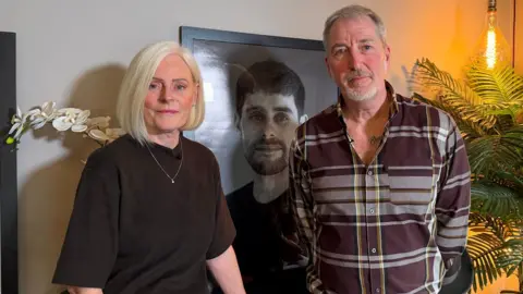 Jim Scott/BBC Lesley and Robin Perkins looking into the camera with a black and white photograph of Dan behind them in a large frame. Lesley is on the left and has a slight smile. She has blonde hair which hangs around her chin and is wearing a black top. Robin is on the right and has grey hair and a goatee. He is wearing a brown, yellow and white checked shirt with the top buttons open revealing a tattoo on his upper chest and has his hands behind his back.