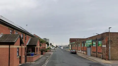 Section of Weelsby Street in Grimsby. There is a single storey factory building on the right and red brick houses on the left