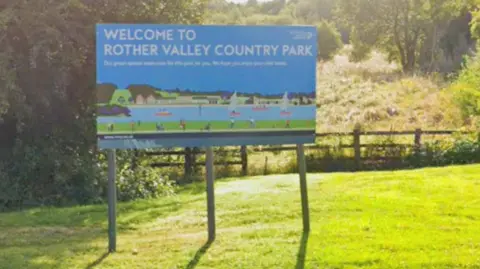 The image shows a large welcome sign for Rother Valley Country Park standing on a grassy area. The sign features illustrations of people sailing and kayaking, along with the text Welcome to Rother Valley Country Park. Behind it is a mix of trees, bushes, and sloping ground.