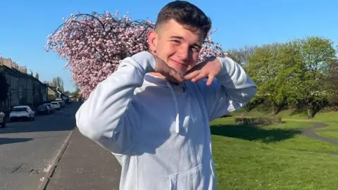 Cole Cooper, a teenage boy. He is wearing a white hoodie and has his hands placed under his chin in a v-shape as if he is framing his face. He is standing on a pavement next to what looks like a park with grass and trees behind him. He was last seen on Cumbernauld Road on 7 May.