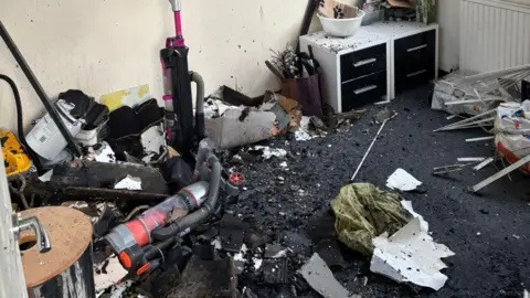 Jess Burnside Black ashes cover the floor of a room, with debris strewn across it. A vacuum cleaner and two sets of drawers are visible against the wall.