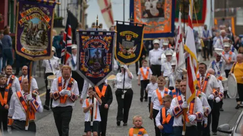 A large group of people holding band flags wearing white shirts and orange sashes.