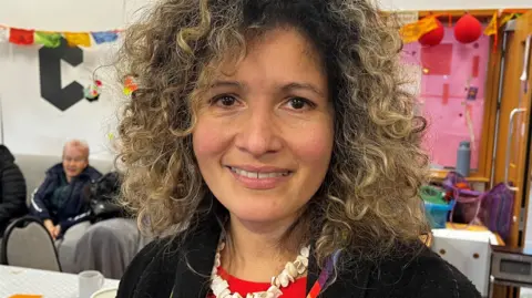 Natali Guevara is standing in a community centre, looking straight to camera. She has shoulder-length light brown curly hair, brown eyes and is wearing a red top and a dark jacket. 