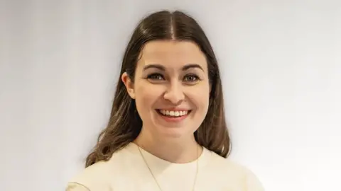 Lauren Troiano standing by a wall, wearing a white top. She has long dark hair and a necklace on, and is smiling.