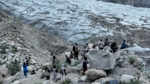 Bahu Trust/Islamic Help A group of people stand and sit among large rocks at the edge of a glacier in a rugged mountain landscape. The glacier stretches across the background, showing layers of blue-white ice streaked with dark sediment and surrounded by steep rocky slopes.
