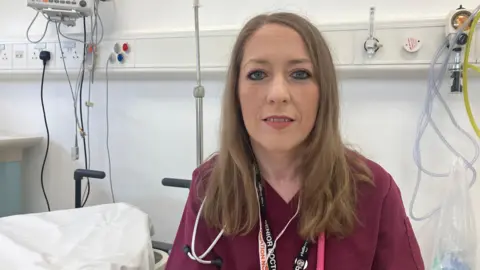 A woman wearing maroon hospital scrubs. She is in a hospital ward and is looking at the camera. She has long brown hair. 