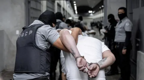 Secretaria de Prensa de la Presidencia/Handout via Reuters Police officers escort alleged members of the Venezuelan gang Tren de Aragua recently deported by the US government to be imprisoned in the Terrorism Confinement Center (CECOT) prison