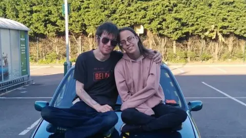 A man and a woman sitting cross legged on a car bonnet looking into the camera. The man has black hair and is wearing sunglasses, jeans and a black T-shirt. The woman has glasses and is wearing trousers and a light brown hooded sweatshirt. 