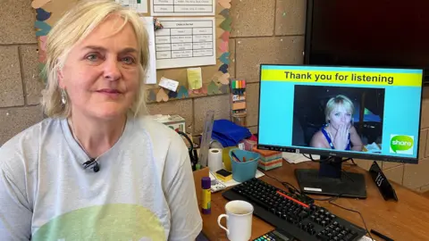 BBC A woman with blonde hair sat at a desk next to a computer which has an image of her daughter on and the words "thanks for listening" above the photo. On the desk are a coffee cup and some stationary, including pens, paper and a ruler.