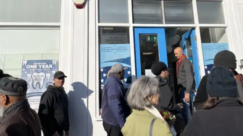 Eight people stood outside the front door to the dental surgery. A man is holding the door open for those entering the building.