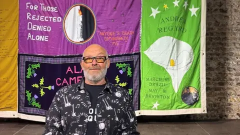 A man wearing a black and white overshirt stood in front of a multi-coloured quilt.