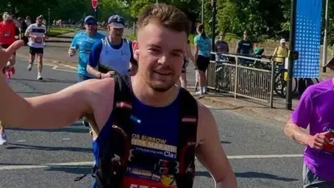 Angus Read A man with brown hair is wearing a running vest and is running alongside other people on a road. He is holding his hand in the air and looks ahead.