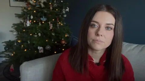 Sarah Drage sites in front of a Christmas tree. She is wearing a red sweater and has long straight black hair.