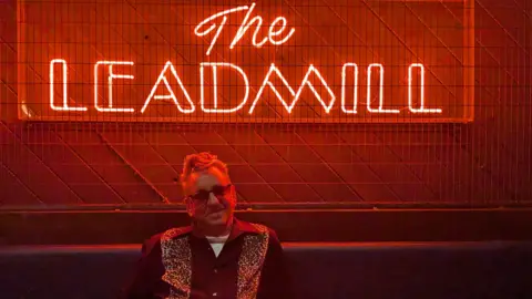 Richard Hawley sat under a red neon light which reads 'The Leadmill'