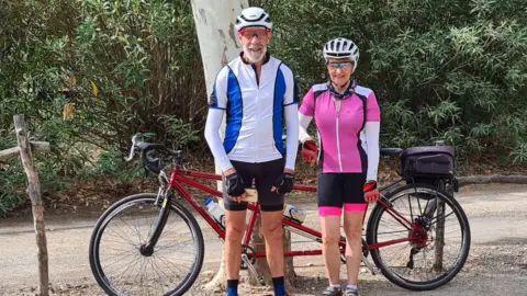 Barry Wilson Barry and Janet stood in front of a red tandem bike which is leant up against a tree in an outdoors area. They are both smiling at the camera. Barry is wearing black cycling shorts, a blue and white long-sleeved cycling top, a white helmet and cycling glasses. Janet is wearing pink and black cycling shorts, a pink and white long-sleeved cycling top, a white helmet and cycling glasses. 