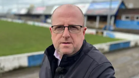Yunus Mulla/BBC Pete Murphy looking into the camera with his mouth slightly open. He is mostly bald with brown and grey short hair on the side of his head. He is wearing rectangular glasses and has stubble over his chin. He is standing in front of a rugby pitch which is out of focus.