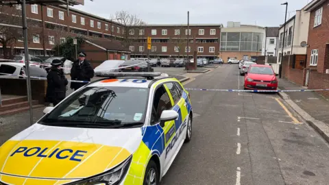 Two police officers in uniform stood by the cordon. A police car is parked to the left of the photo.