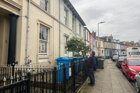 A man stands with his hand on metal railings outside a colourful terrace of homes, painted in shades of cream, green, white, pink, blue and yellow. Behind him, the road sweeps round to the right. Cars are parked by the side of the road. Blue and black bins are lined up outside the homes. The sky is grey and cloudy.