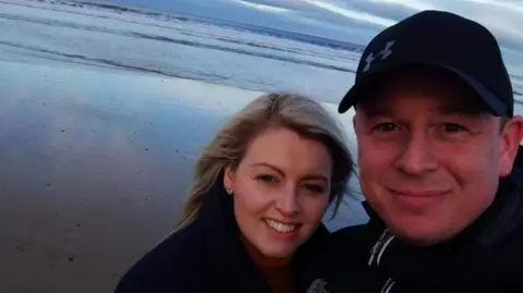Samantha Jones A selfie of Samantha and Trevor. They're standing on a beach and smiling into the camera. Samantha has her hair down and Trevor is wearing a black cap.