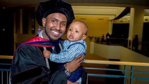 Aj Awer Aj Awer is dressed in cap and gown, posing for a photo with his son
