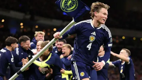 Getty Images Scott McTominay celebrating with the corner flag. He is wearing a navy blue Scotland shirt and shorts.