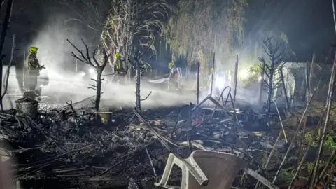 Maldon Fire Station Another view of the destroyed gardens as firefighters hose down the affected area. Rubble is littered across the scene and burnt trees can be seen.