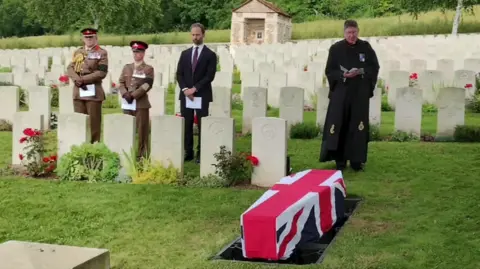 Sue Kruk A funeral service at a British war cemetery in France, a coffin with a British flag draped across it is being lowered into the ground and a vicar is reading beside military personnel 