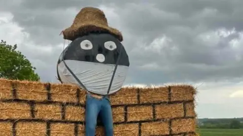 A block of straw bales, with painted lines to look like bricks and perched on top a black and white round plastic coloured bale, with a eyes and a nose with its legs - stuffed blue jeans - dangling below
