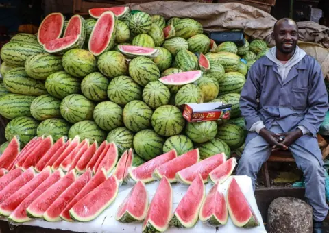 JAMES WAKIBIA / SOPA IMAGES / LIGHTROCKET / GETTY IMAGES A man sits next to piles of dozens of large melons on his stall.