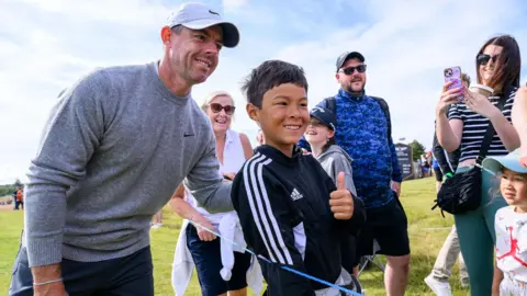 PA Media Rory McIlroy, who is wearing a grey round neck jumper and a silver baseball cap poses with a smiling young golf fan. The boy is wearing a tracksuit top and is giving a thumbs up to the camera