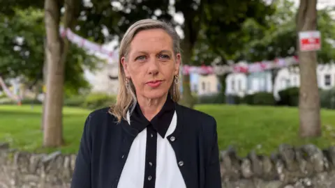 A woman in a white shirt and black cardigan. She has blonde hair and stands in front of green grass.