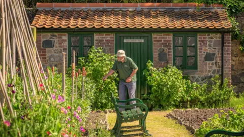 Saumarez Park The image shows a man gardening in front of a small brick building with a green wooden door and green window frames. The man is wearing a hat and is looking to the left as he tends a section of the garden, which has pink and red flowers.