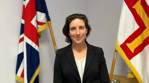 Deputy Lindsay de Sausmarez - A woman with black hair at shoulder length. She is wearing a black jacket over a white patterned blouse. She is standing in front of a Guernsey and British flag. 