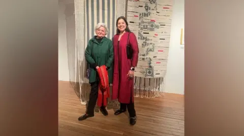 Manmeet Walia Two women stand inside a gallery. The room has plain, light-coloured walls and a polished wooden floor. Behind them hangs a large textile artwork. The woman on the left is wearing a dark green coat over black trousers and black shoes. The woman on the right is wearing a long, deep red outfit over dark trousers with black shoes. 