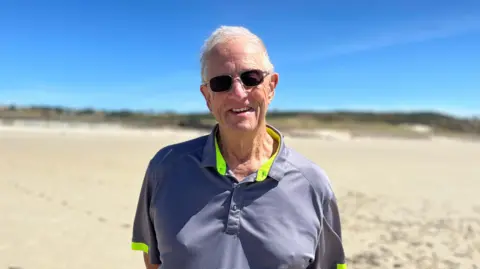 Michael is wearing sunglasses and a grey polo shirt stands on a wide sandy beach with hands behind their back. Sand dunes and vegetation are visible in the distance beneath a clear blue sky.