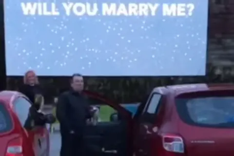 Karen Hall/Facebook A man stands next to a red car, with a cinema screen behind him which reads 'WILL YOU MARRY ME?'. 