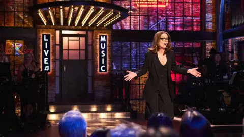 Tina Fey on the set of Saturday Night Live UK with a sign saying live music behind her - a band can be seen on the stage, and audience members are in front of her